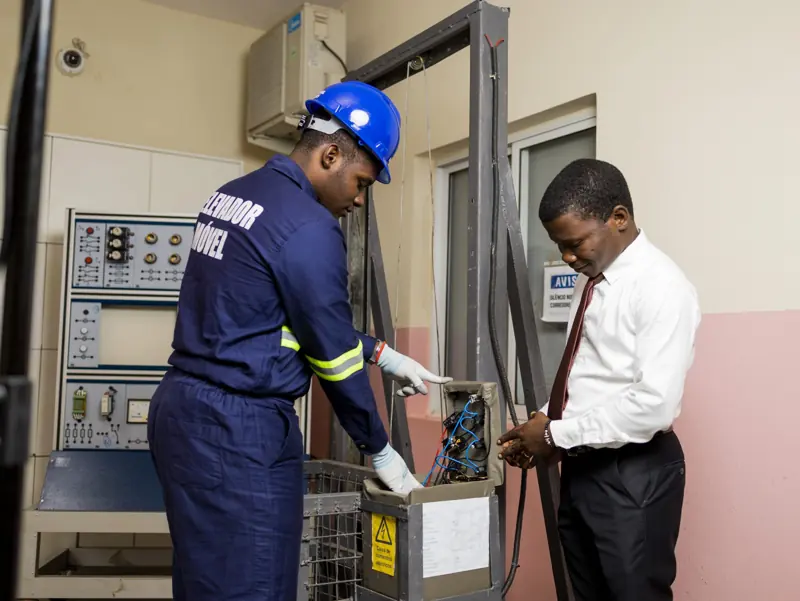 Aluno de Electromecânica a trabalhar num painel de elevador, com um professor ao lado